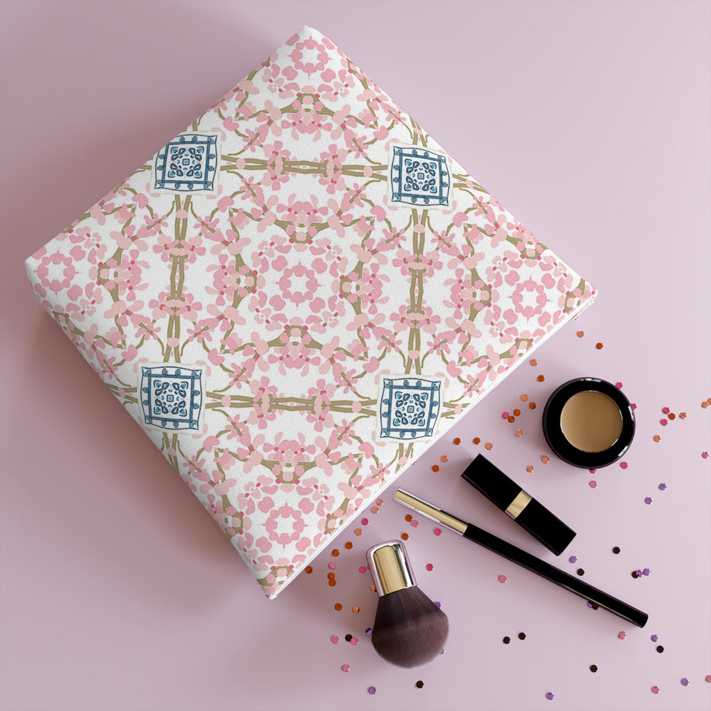 Cosmetic bag with white zipper and design of pink flowers in a circular pattern with blue and white motif at the center of brown twigs crossing on a white background and laying on it's side with make up contents and confetti coming out of it.