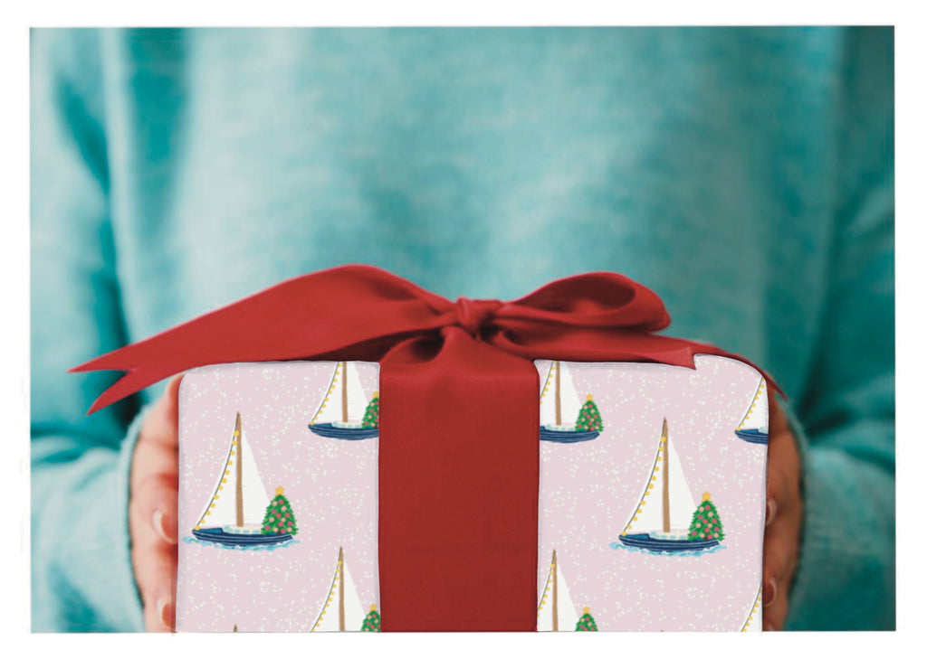 Gift with wrapping paper of Sailboats with lights and christmas tree on it on a light pink background and a red bow.