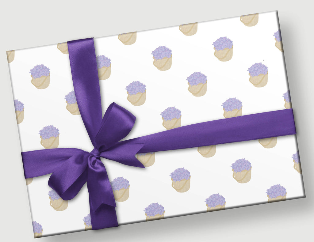 This is a photo of a gift with purple ribbon and bow.  The gift wrap is an arrangement of the same motif - hand designed lavender colored hydrangeas in a Nantucket basket and they are scattered on a white background. 